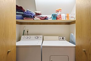 Une buanderie équipée de deux machines à laver et deux sèche-linge superposés, une étagère avec des serviettes pliées et du détergent à linge sur l’étagère supérieure.