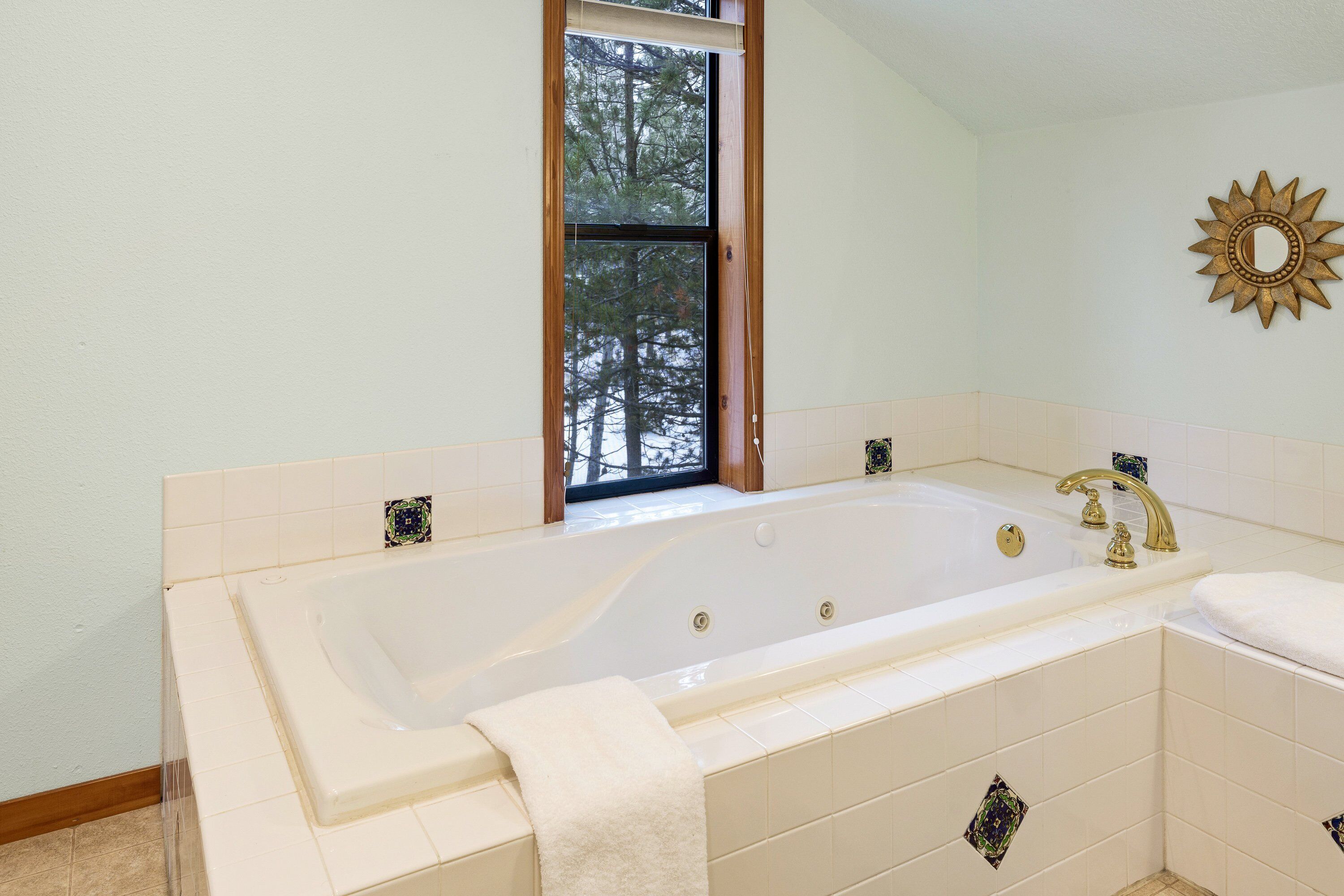 A bathroom with a large bathtub, a window with a view of trees, and a decorative sun-shaped mirror on the wall.