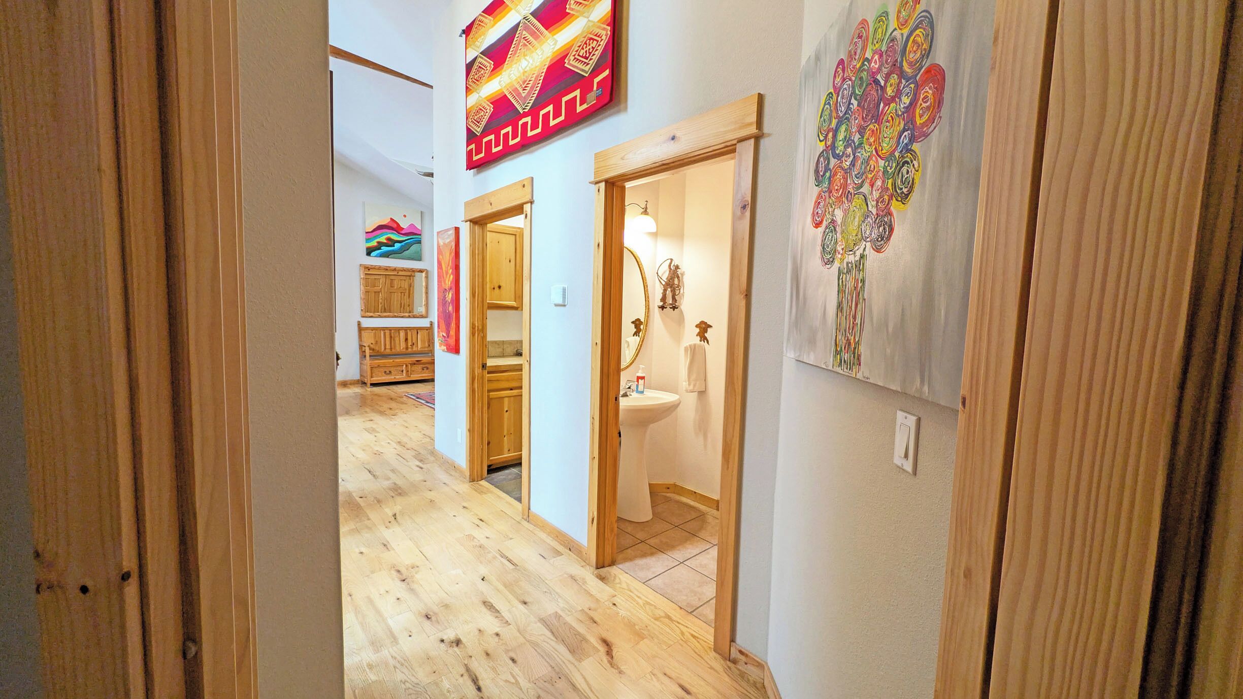 A hallway with wooden flooring, a bathroom with a sink and mirror, and artwork on the walls.