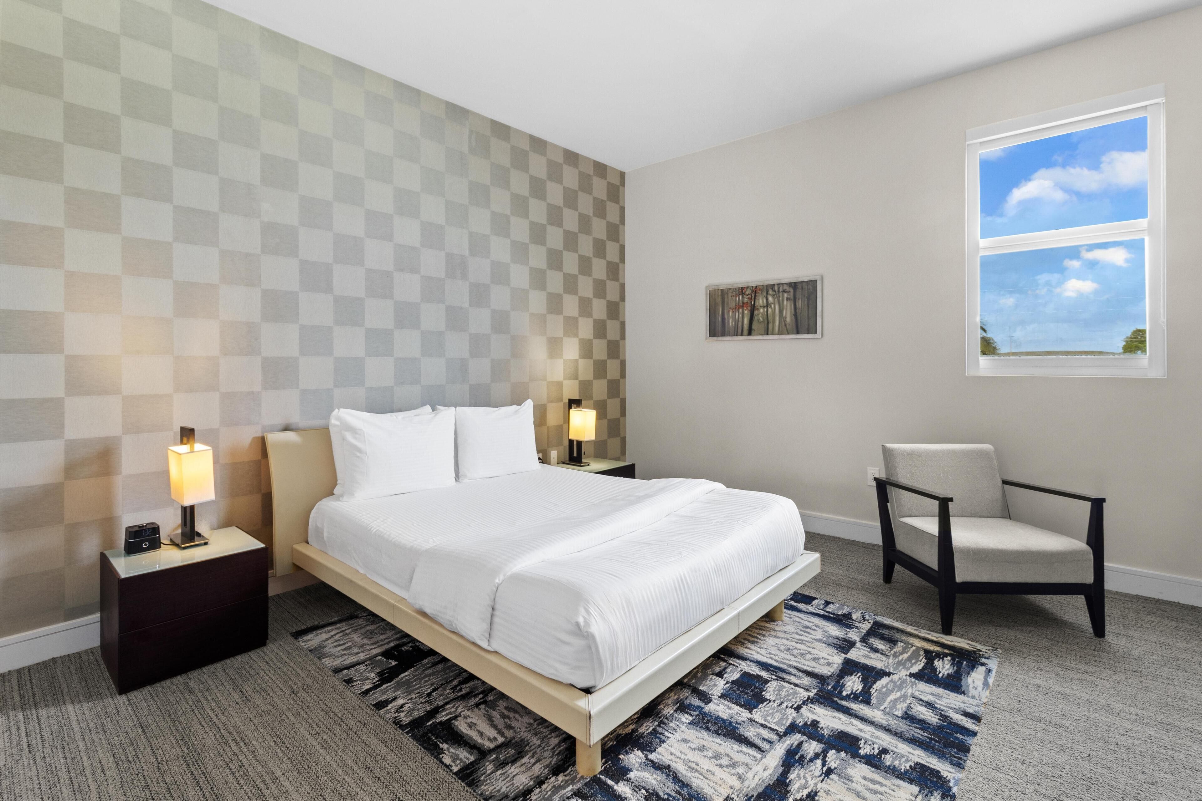 A hotel room with a checkered wall, a bed with white linens, two bedside lamps, a chair, and a window with a view.