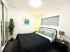 Un dormitorio moderno con una cama negra, una mesita de noche con lámpara, una ventana con persianas, una planta en maceta y un cuadro en la pared.