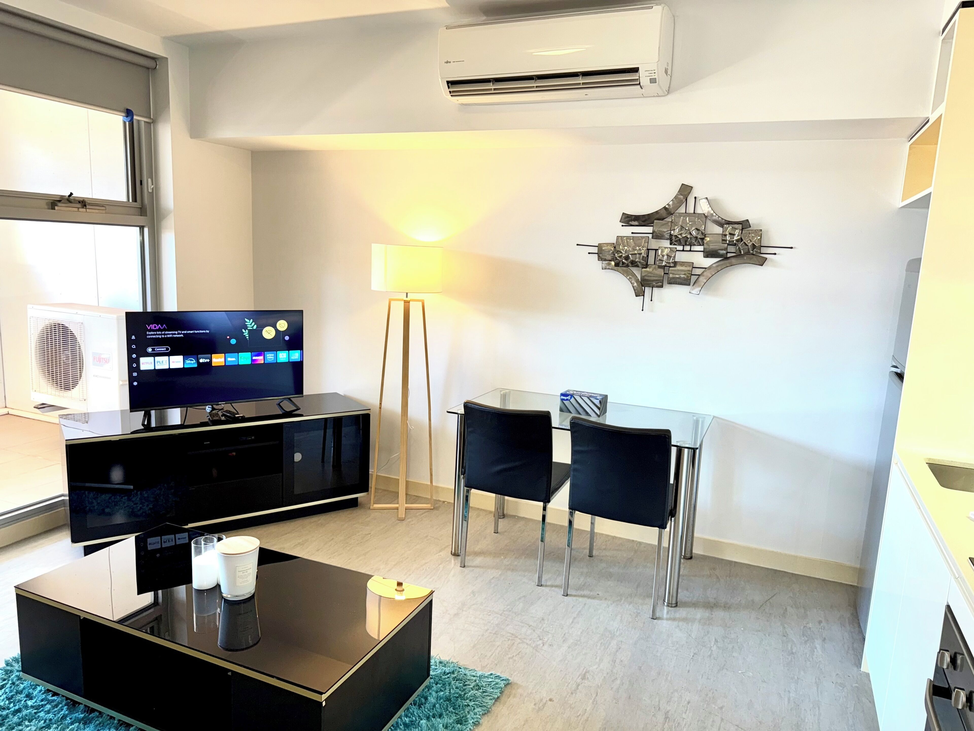 A modern living room with a flat-screen TV, a glass coffee table, a lamp, and a dining area with chairs.