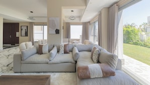 A spacious living room with a large sectional sofa, a wooden coffee table, and a view of a well-maintained garden through large windows. - ALSAWAH Spacious Family Villa in Palm Hills Sheikh Zayed (6th of October City)
