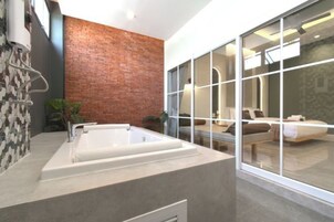 Un baño moderno con una bañera grande, una pared de acento de ladrillo y un ventanal con vistas a un dormitorio.