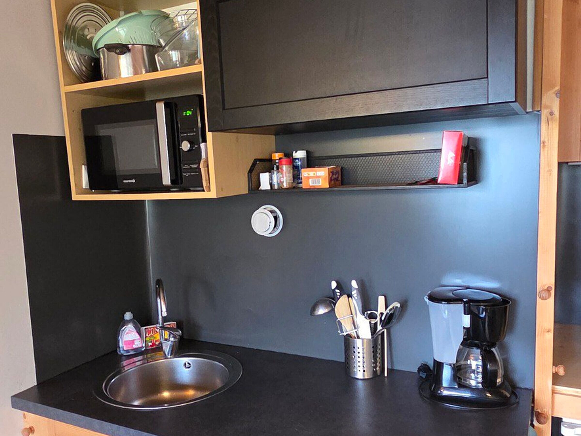 A compact kitchen with a black countertop, a stainless steel sink, a microwave, a coffee maker, and a utensil holder.