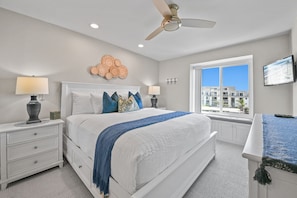 A bedroom with a bed, bedside tables, a ceiling fan, a wall-mounted TV, and a window with a view of buildings.
