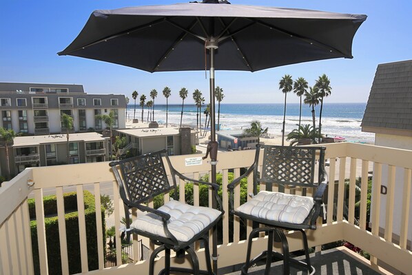 A balcony with two chairs and an umbrella, overlooking a beach with palm trees and a pier.