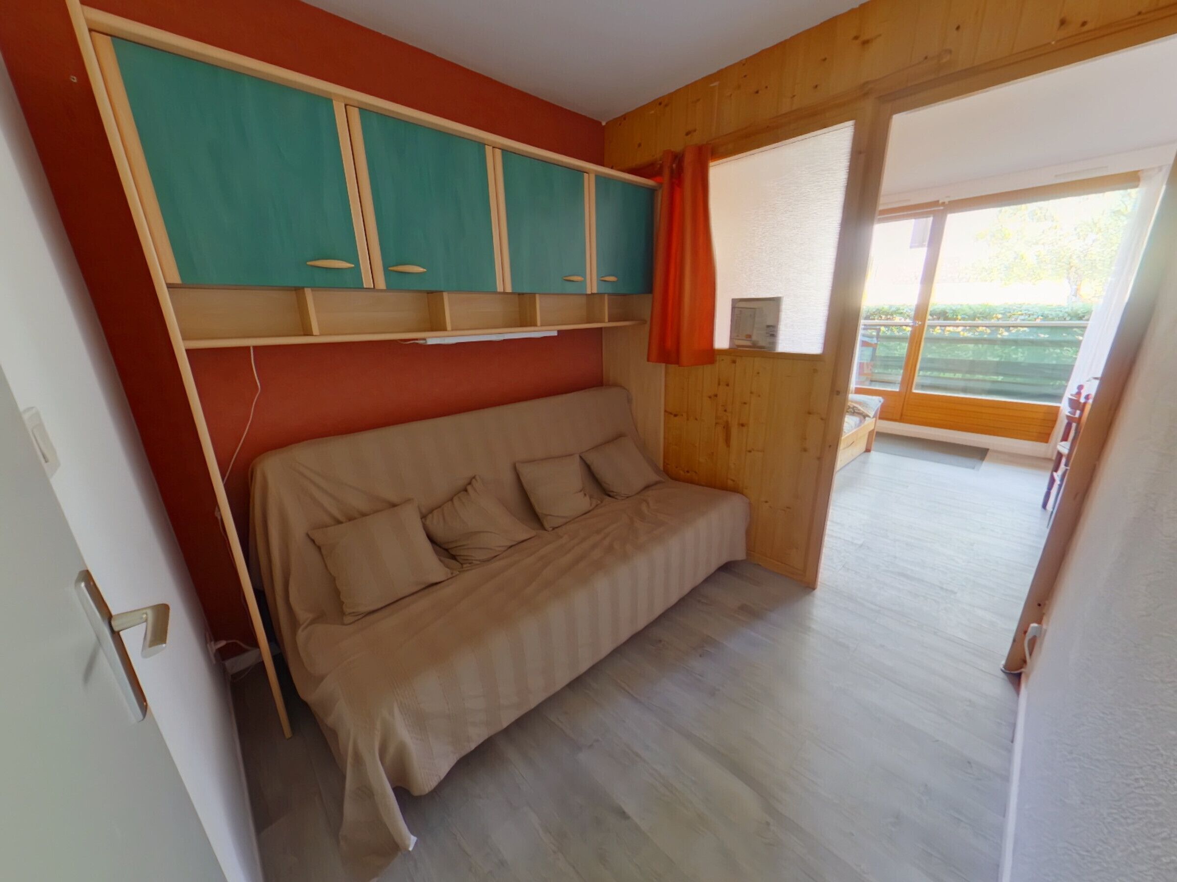 A room with a sofa bed, wooden panel walls, a window with curtains, and a view of greenery outside.