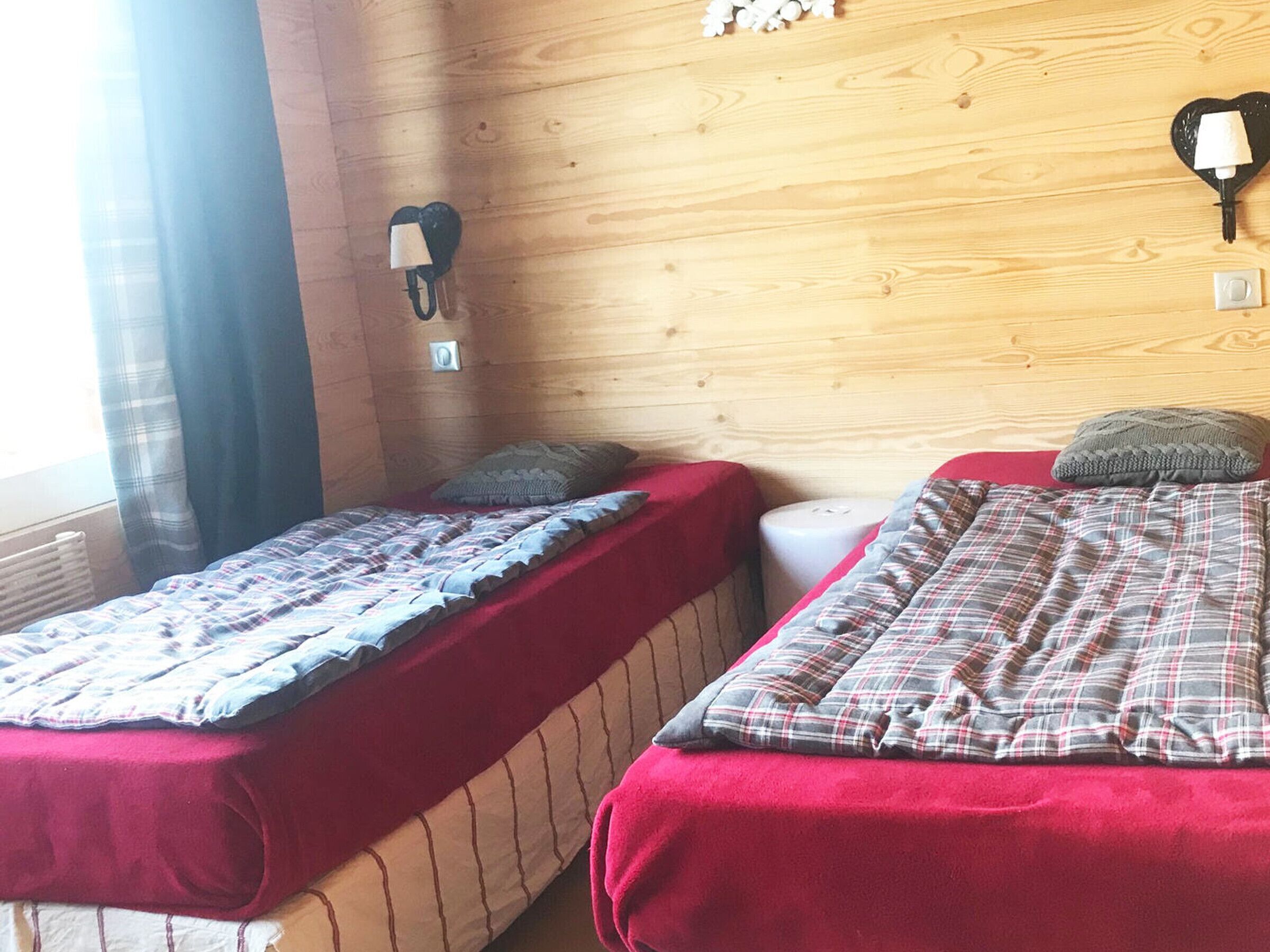 A room with two beds, each with a red blanket and a plaid duvet. There are wall-mounted lamps and a small heater on the wall.