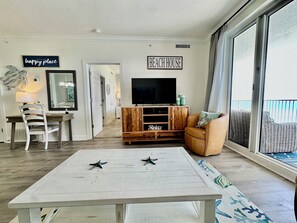A living room with a white coffee table, a television on a wooden stand, a brown leather sofa, and a large window with a view.