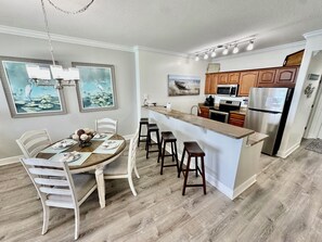 A modern kitchen with a central island, wooden cabinets, stainless steel appliances, and a dining area with white chairs and a table set for four.