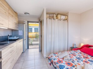 Quarto compacto com cozinha compacta, uma cama com colcha estampada em flores coloridas e vista para um prédio amarelo através de uma porta.