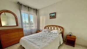 Een slaapkamer met een houten bed, een houten dressoir, een nachtkastje met een lamp, en een raam met dunne gordijnen.