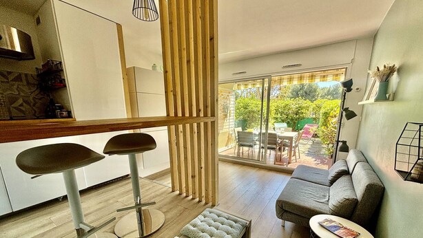 A modern living room with a sofa, two bar stools, a coffee table, and a bench. There is a large sliding glass door leading to an outdoor patio area with a table and chairs.