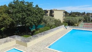 A rectangular swimming pool with a stone deck and white railing, surrounded by greenery and buildings.