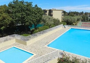 Una piscina rectangular con terraza de piedra y barandilla blanca, rodeada de vegetación y edificios.