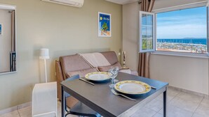 A dining area with a table set for two, a window with a view of the sea, and an air conditioning unit on the wall. - Air-Conditioned Apartment in Le Lavandou Center, Near Beach (Le Lavandou)