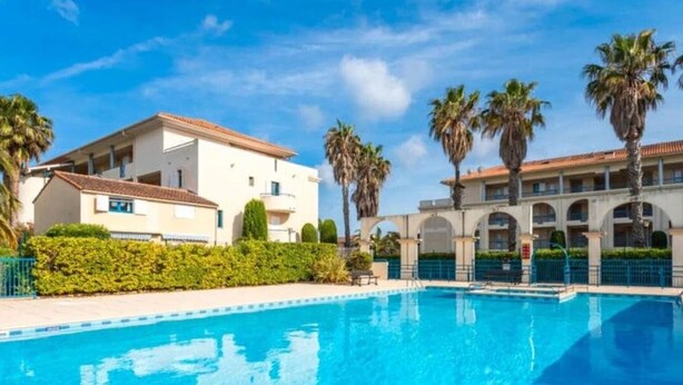 A swimming pool with a clear blue water surface, surrounded by a low hedge and a building with a tiled roof.