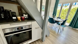 A modern kitchen with a built-in oven, a coffee maker, and a kettle. There is a dining area with a table and chairs, and a view of greenery through large windows.