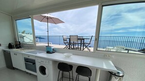 Une cuisine compacte équipée d’un lave-linge, d’un grille-pain et d’un évier. Un balcon avec une table et des chaises offre une vue sur la mer.
