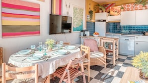 Un coin repas avec un sol à carreaux, une table dressée pour quatre personnes et, au fond, une cuisine dont le mur derrière l’évier est recouvert de carreaux bleus.