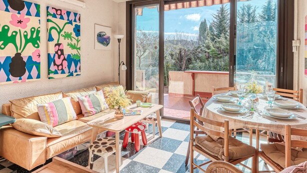 Un salon chaleureux avec un canapé, une table basse et une table à manger dressée pour un repas. Une grande fenêtre offre une vue sur les arbres et un ciel dégagé.