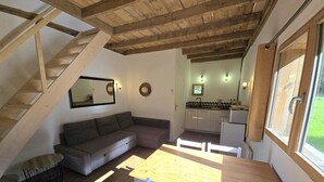 A cozy loft-style living space with a wooden staircase, a gray sectional sofa, a wooden dining table with chairs, a kitchen area with a refrigerator, and a large window.