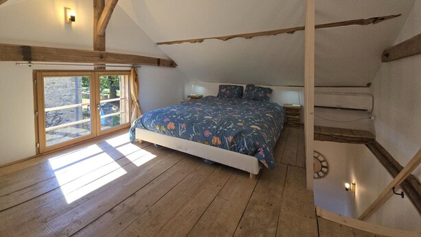 A bedroom with a sloped ceiling, wooden beams, a large bed with floral bedding, a bedside table, a wall-mounted clock, and a sliding glass door leading to an outdoor area.