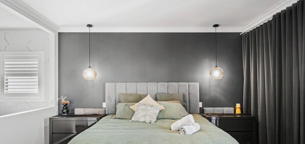Un dormitorio moderno con un cabecero tapizado en gris, dos mesitas de noche, una cama con colcha estampada en verde y dos lámparas colgantes.