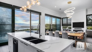 A modern kitchen with a large island, a dining area with a view, and a living space with a flat-screen TV. - Surfside - Beachfront Deluxe, Pool, Spa & BBQ (Catherine Hill Bay)