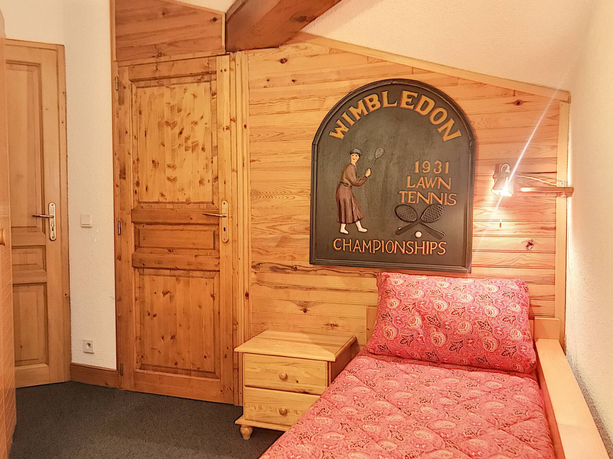 Un dormitorio con un armario de madera, una cama con cubrecama estampado y un cartel en la pared sobre el Campeonato de Wimbledon de Lawn Tenis 1931.