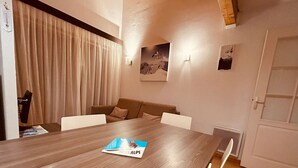 A modern living room with a wooden dining table, a beige sofa, and a white armchair. There are two wall-mounted light fixtures and a door with a glass panel. - Bright 3-room apartment with parking in Serre Chevalier (Serre Chevalier Chantemerle)