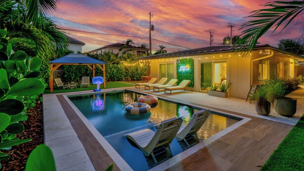 Una piscina con sillas de descanso y una zona de estar cubierta, rodeada de vegetación y un cielo al atardecer.