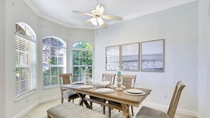 A dining room with a wooden table set for four, large windows with blinds, a ceiling fan, and framed artwork on the wall. - Dolphin Dreams | Private Pool | Naples Park Retreat (Naples)