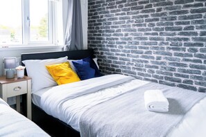 En pænt redt seng med hvide, gule og blå puder, et gråt tæppe og et sammenrullet hvidt håndklæde på et trænatbord.