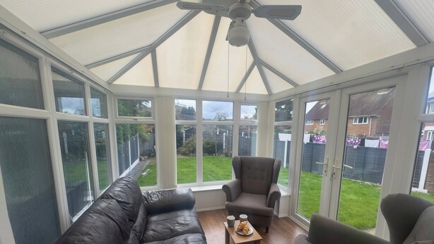 Ein Wintergarten mit Deckenventilator, einem schwarzen Ledersofa, einem grauen Sessel und einem kleinen Tisch mit einem Croissant und zwei Tassen.