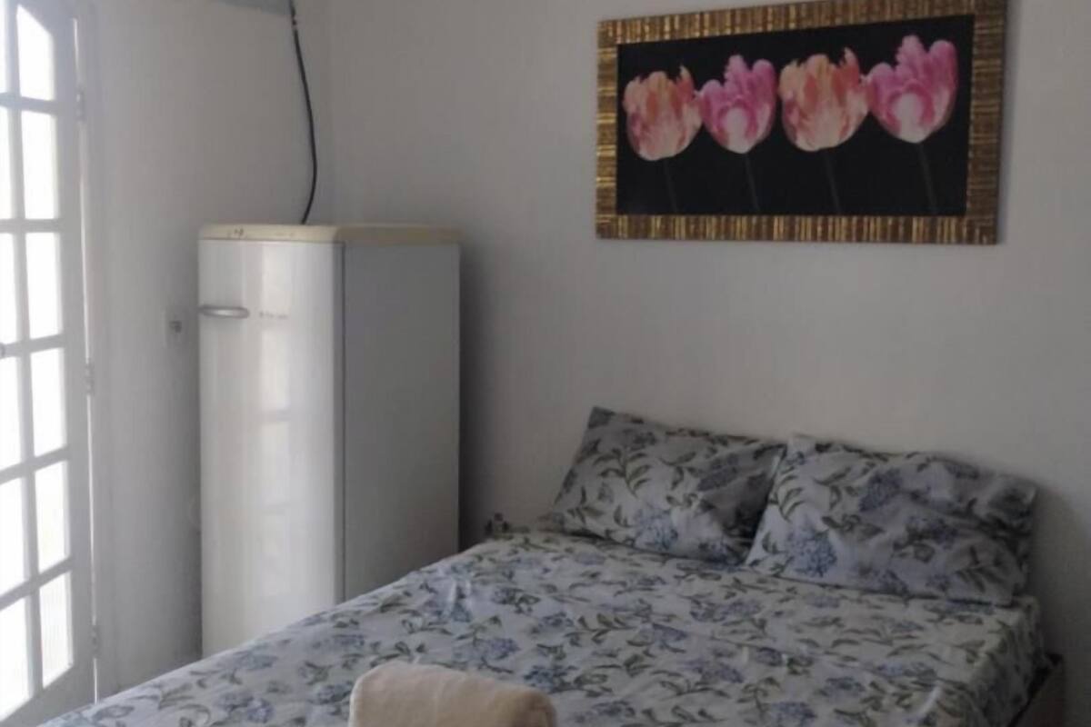 A bedroom with a bed, a wall-mounted air conditioner, a window, and a floral painting.