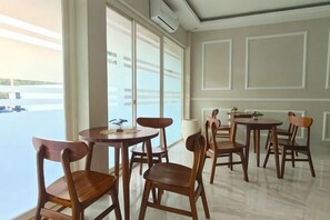 A dining area with round tables and wooden chairs, large windows with blinds, and a view of a body of water. - El Nipa Hotel Labuan Bajo (Wae Kelambu)