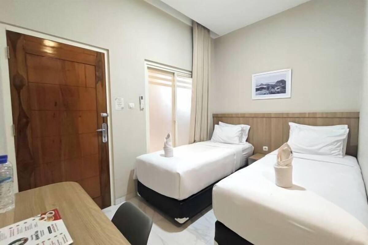 Ein Hotelzimmer mit zwei Betten, einer Holztür, einem Fenster mit Vorhängen und einem gerahmten Bild an der Wand.