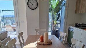 A modern kitchen with a wooden table, white chairs, a clock, and a view of a balcony with plants. - Residencial Breeze 1A by Bookaparadise (Finestrat)