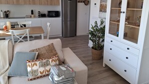 A modern kitchen with white cabinets, a stainless steel refrigerator, and a microwave. There is a dining table with chairs and a sofa with decorative pillows. A cabinet with glass doors is visible in the foreground. - Residencial Breeze 1A by Bookaparadise (Finestrat)