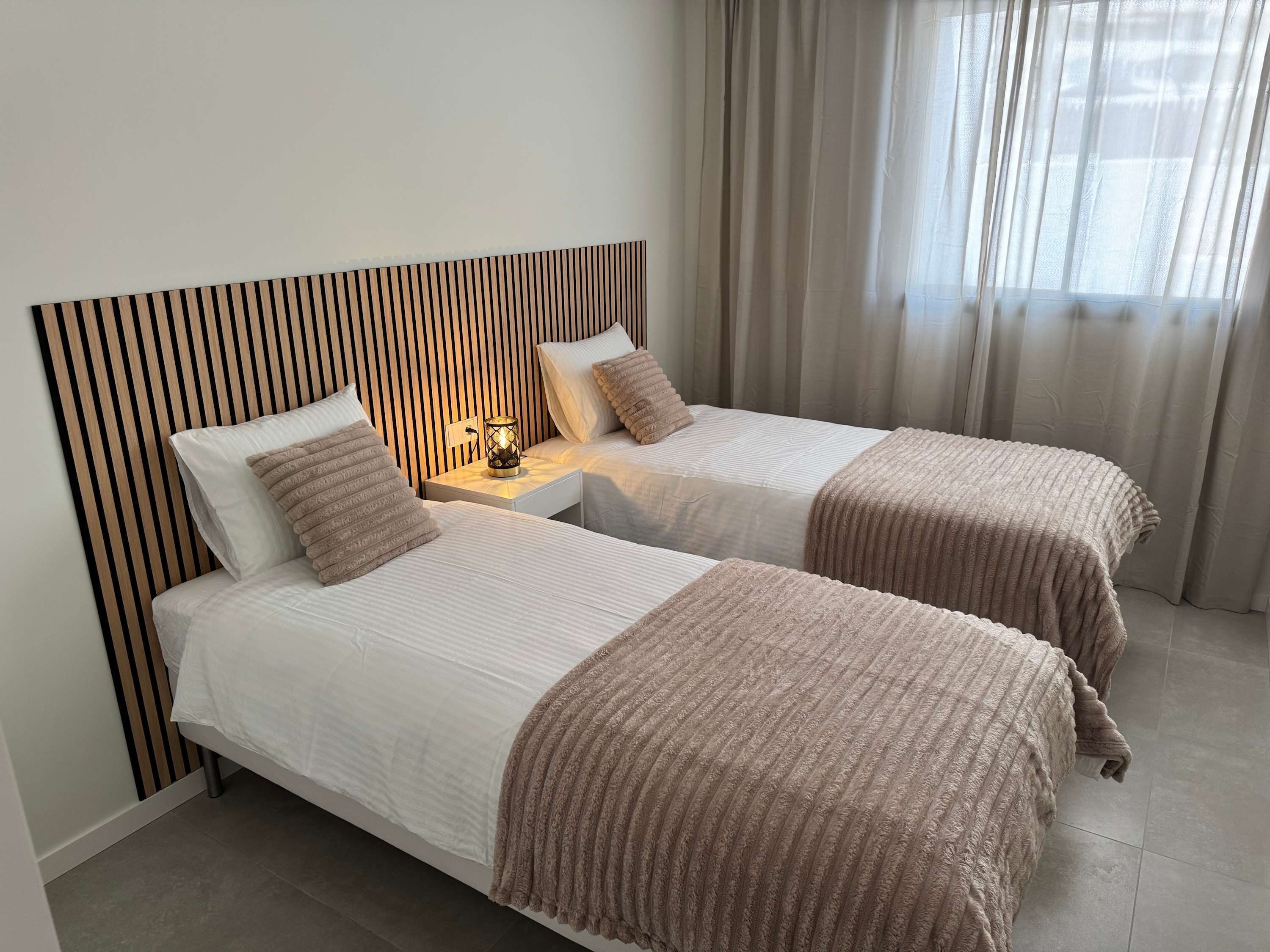 Une chambre d’hôtel avec deux lits, une table de chevet avec une lampe, une tête de lit en bois et une fenêtre avec des rideaux.