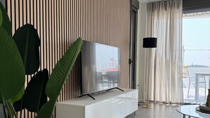 A modern living room with a TV on a white cabinet, a large plant, and a view of the sea through a window. - Resort Seascape Villa 8 by Bookaparadise (Finestrat)