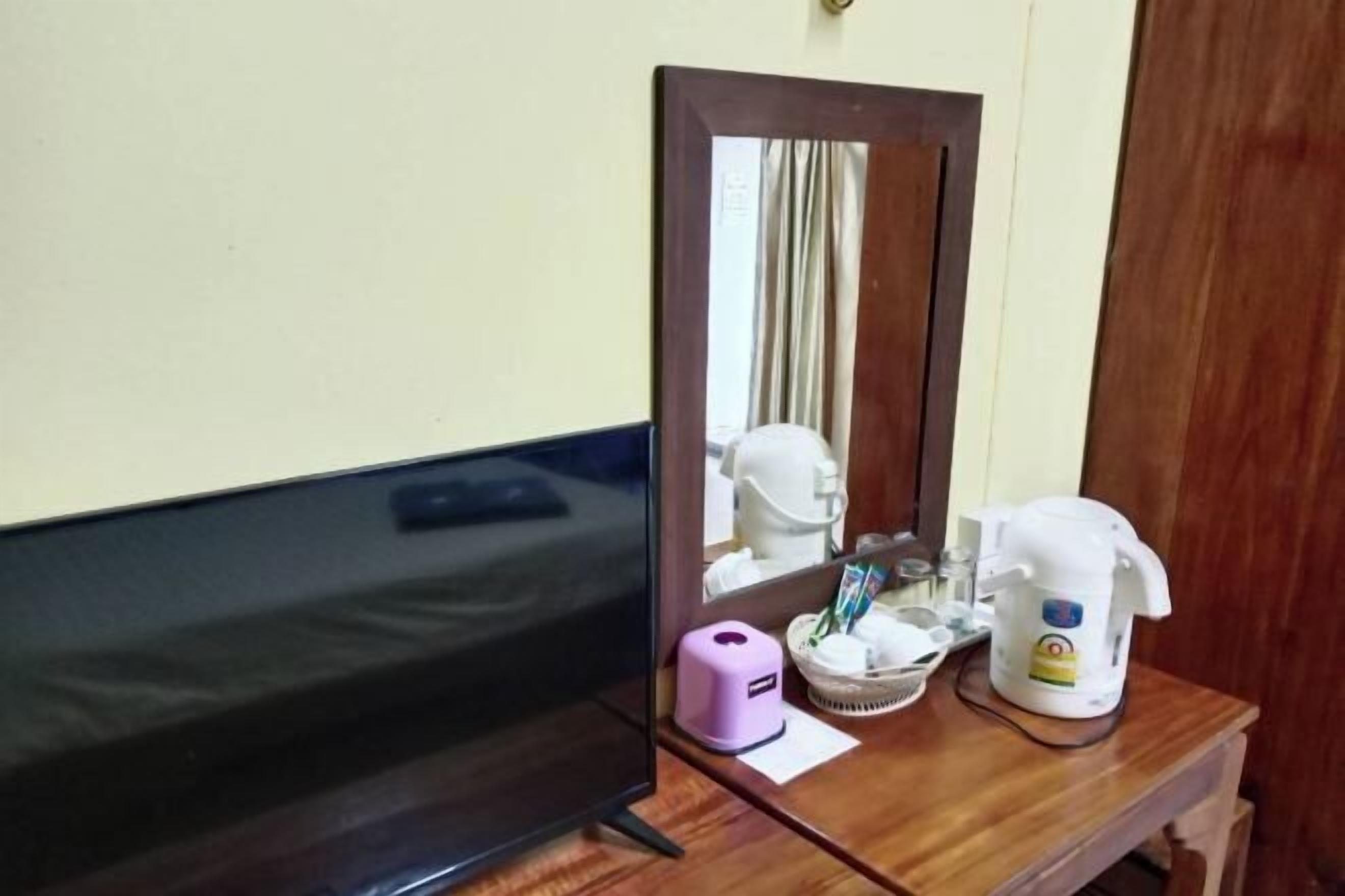 A wooden desk with a television, a mirror, a purple dispenser, a basket, and a white kettle. - Peamsuk Sweet Prachin Buri (Prachinburi)