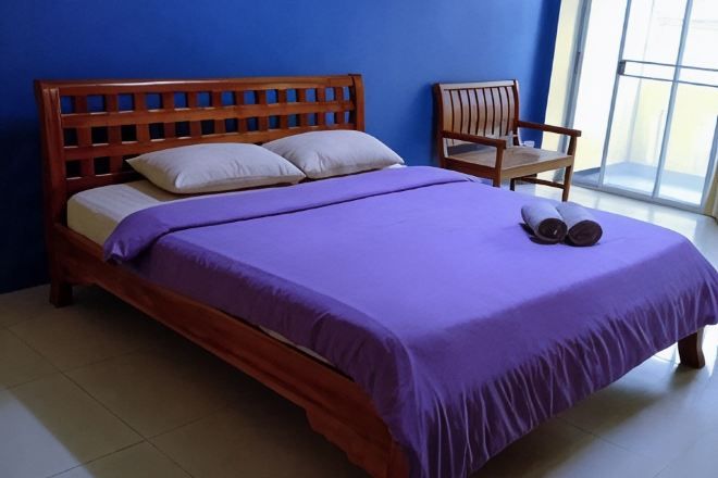 Una cama con cabecera de madera, colcha morada y dos almohadas. Hay una silla al lado de la cama.