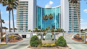 A modern hotel with a curved glass facade, palm trees, and a central sculpture. - NEW! 3.5 bedroom 3.5 bath condo in Orange Beach, walking distance to Flora-Bama! (Orange Beach)