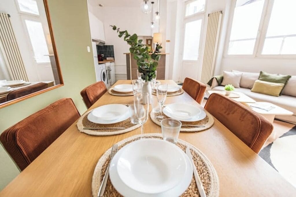 Uma área de refeições com mesa de madeira posta para quatro pessoas, rodeada por cadeiras estofadas em tons de castanho.