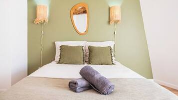A bed with a white mattress, two green pillows, and a rolled-up gray towel on a beige bedspread. There are two wall-mounted lamps with woven shades on either side of a mirror above the bed.