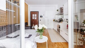 A hotel room with a bed, a small table with a vase of flowers, a kitchen area with cabinets and appliances, and a door with a notice.