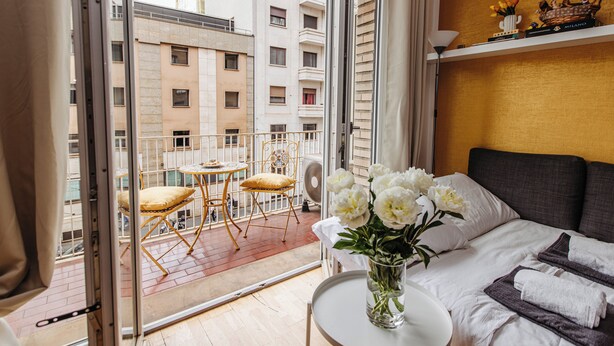 A hotel room with a balcony, a bed with pillows and towels, a small table with a vase of flowers, and a view of the cityscape.
